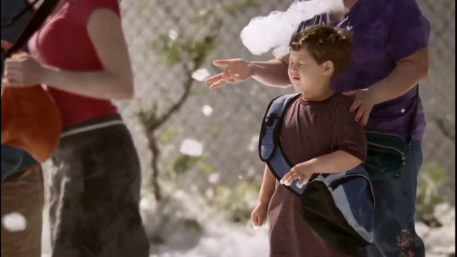 A young boy with a backpack looks up in wonder as foam falls around him, while an adult woman stands beside him, outdoors.