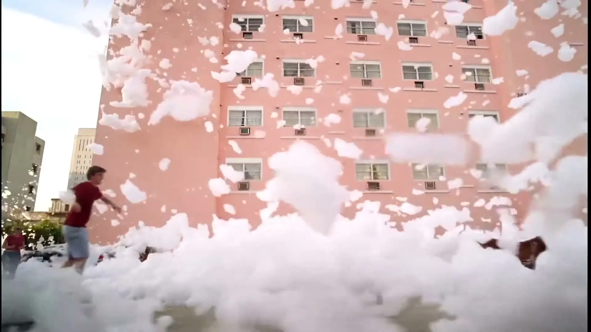 A young man in a red shirt and shorts plays in a street filled with foam, with a pink building in the background.