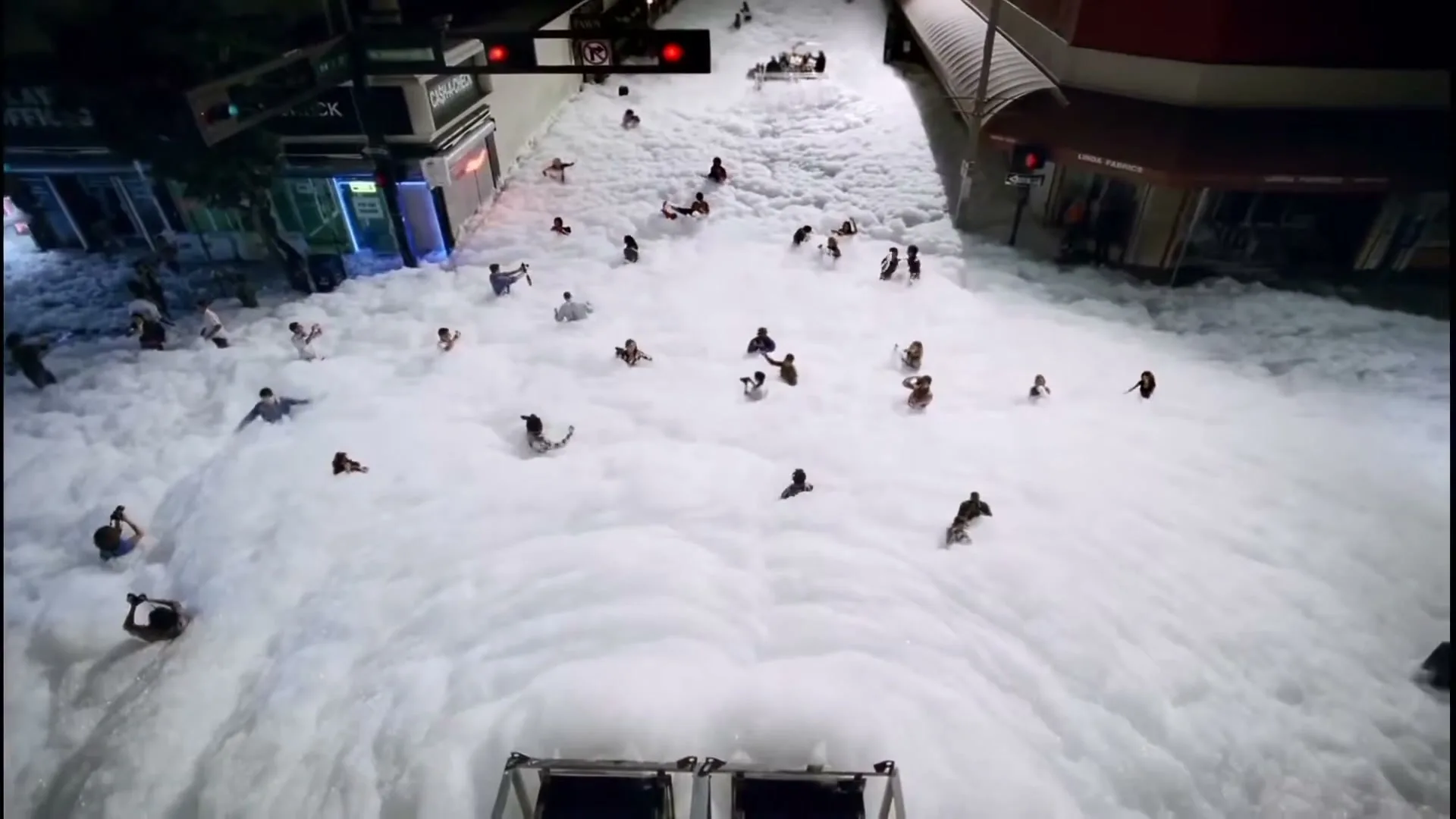 A high-angle shot captures a bustling city street at night, completely submerged in a sea of white foam, with numerous people wading and playing in it.
