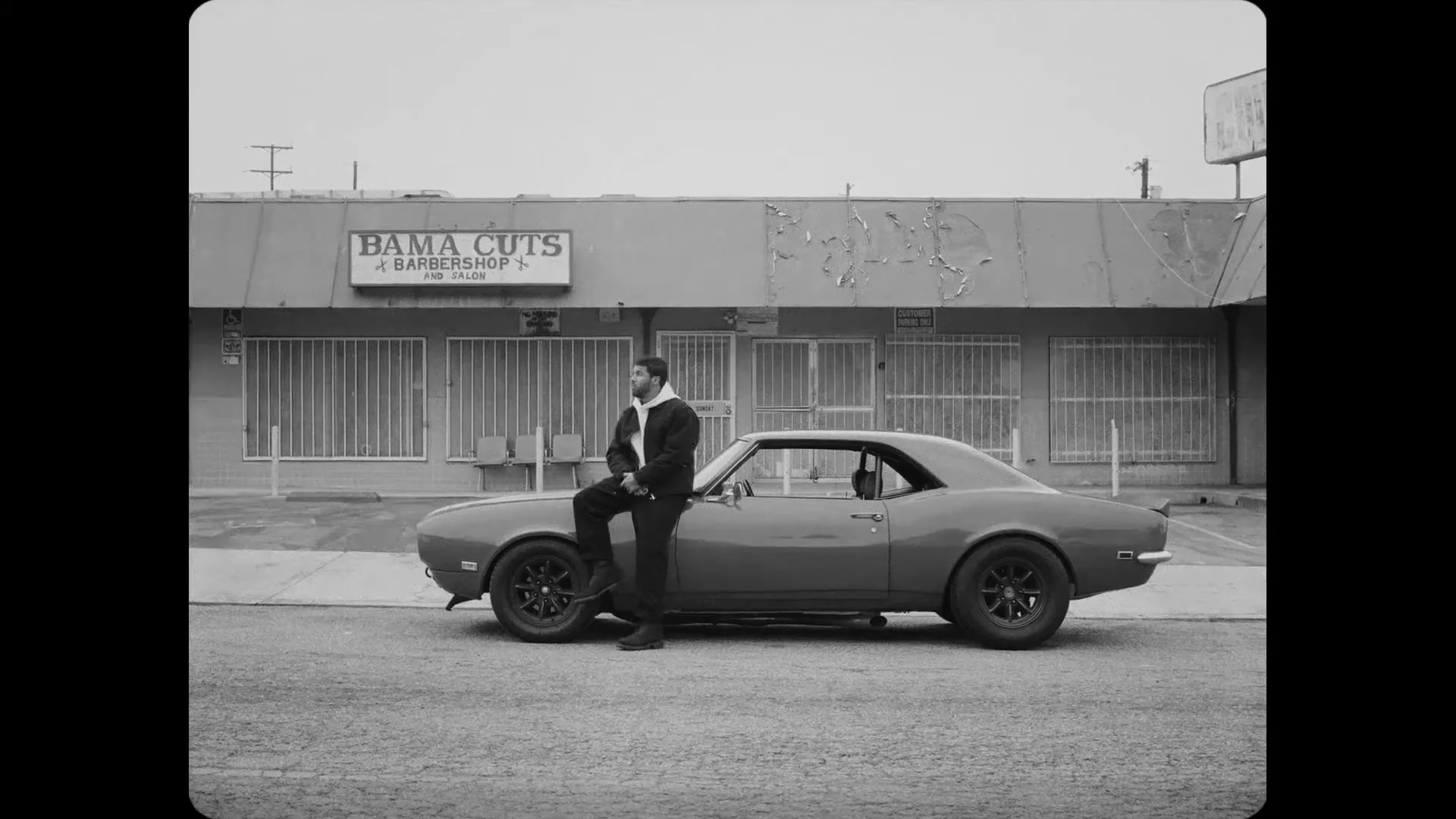 A man in a hoodie and dark pants leans against a classic car parked on a street in front of a barbershop.