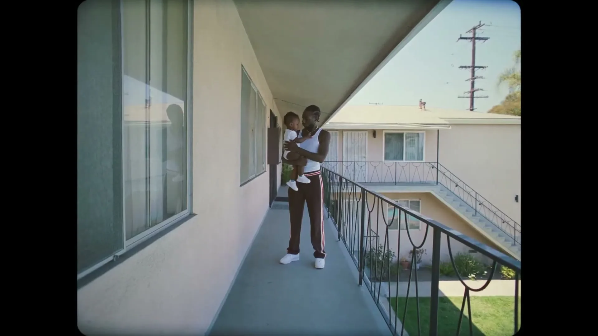 A young Black man in a white tank top and red track pants holds a baby on a balcony overlooking an apartment complex on a sunny day.