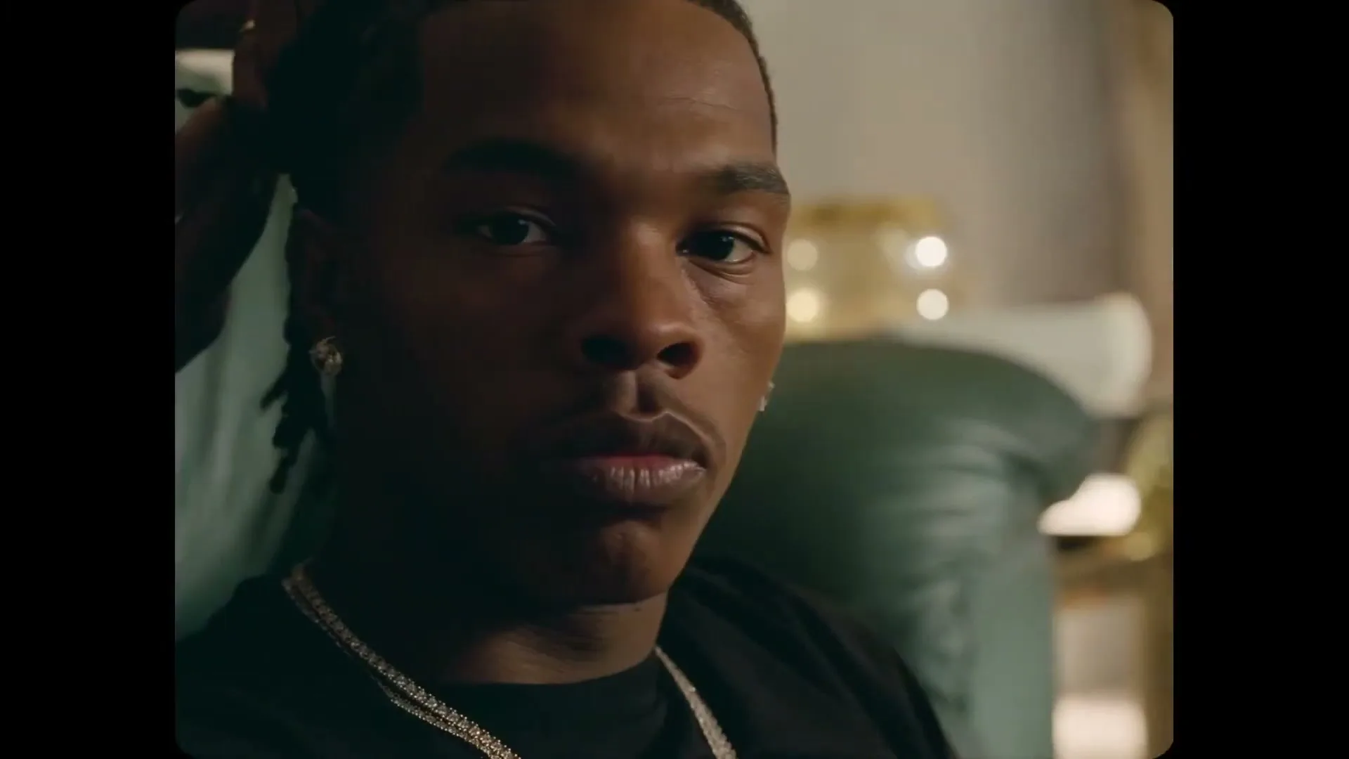 Lil Baby, a young Black man with dreadlocks and diamond earrings, looks directly at the camera while someone touches his hair in an interior setting.