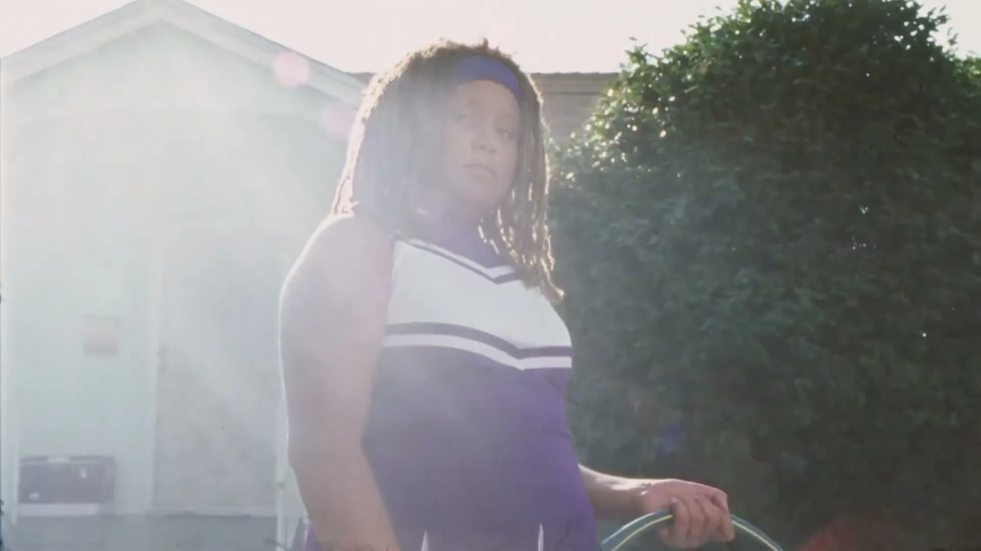 A young person with dreadlocks and a purple and white cheerleading uniform stands outdoors, holding a hose, looking directly at the camera.