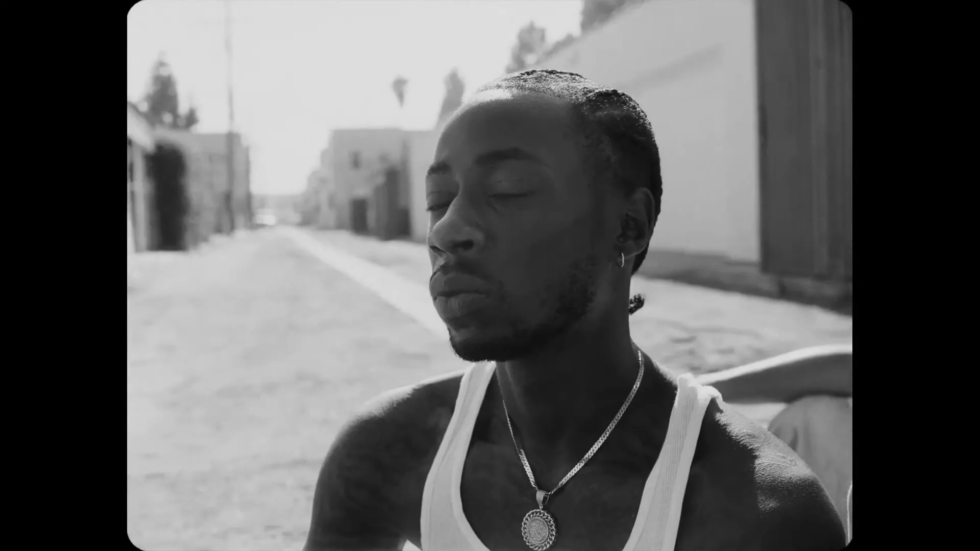 A Black man with braided hair and a white tank top looks around with a neutral expression on a sunny day in a residential alleyway.