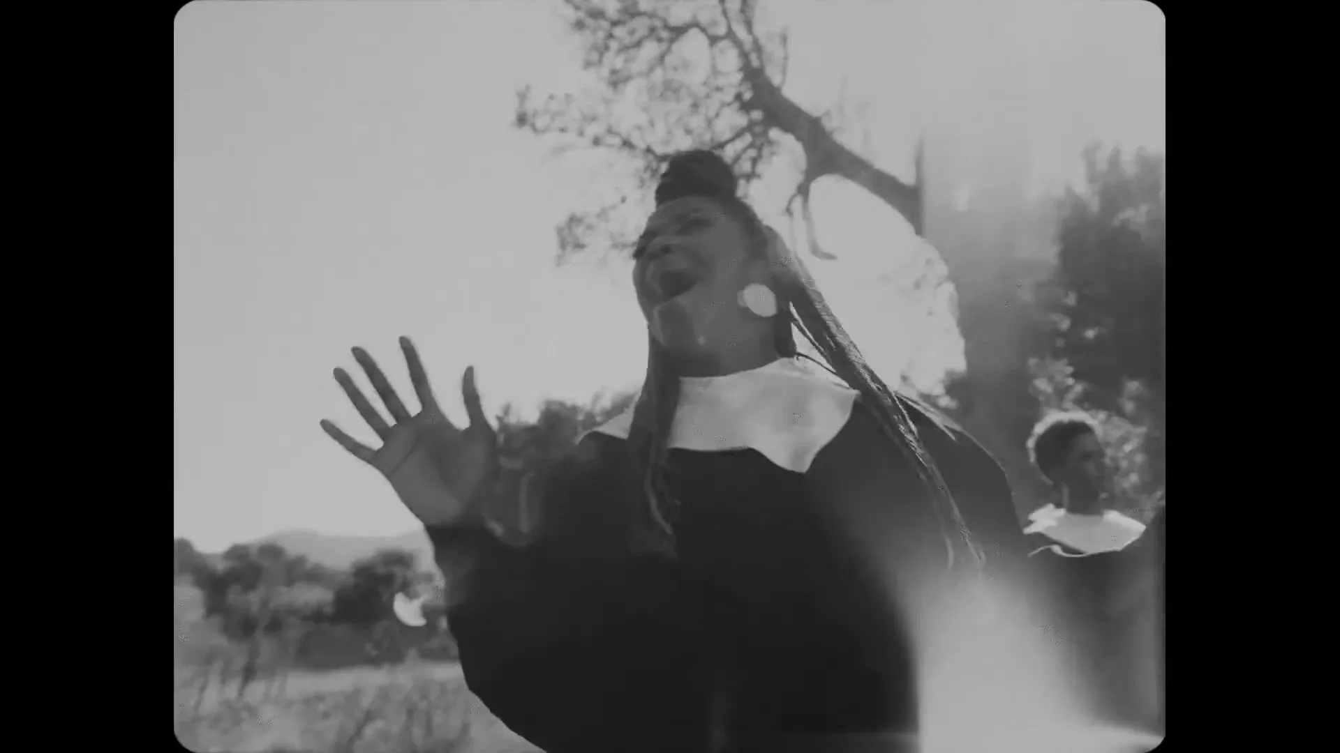 A Black woman with dreadlocks, wearing a nun's habit, sings with her hands raised in a sun-drenched outdoor setting, with a tree in the background.