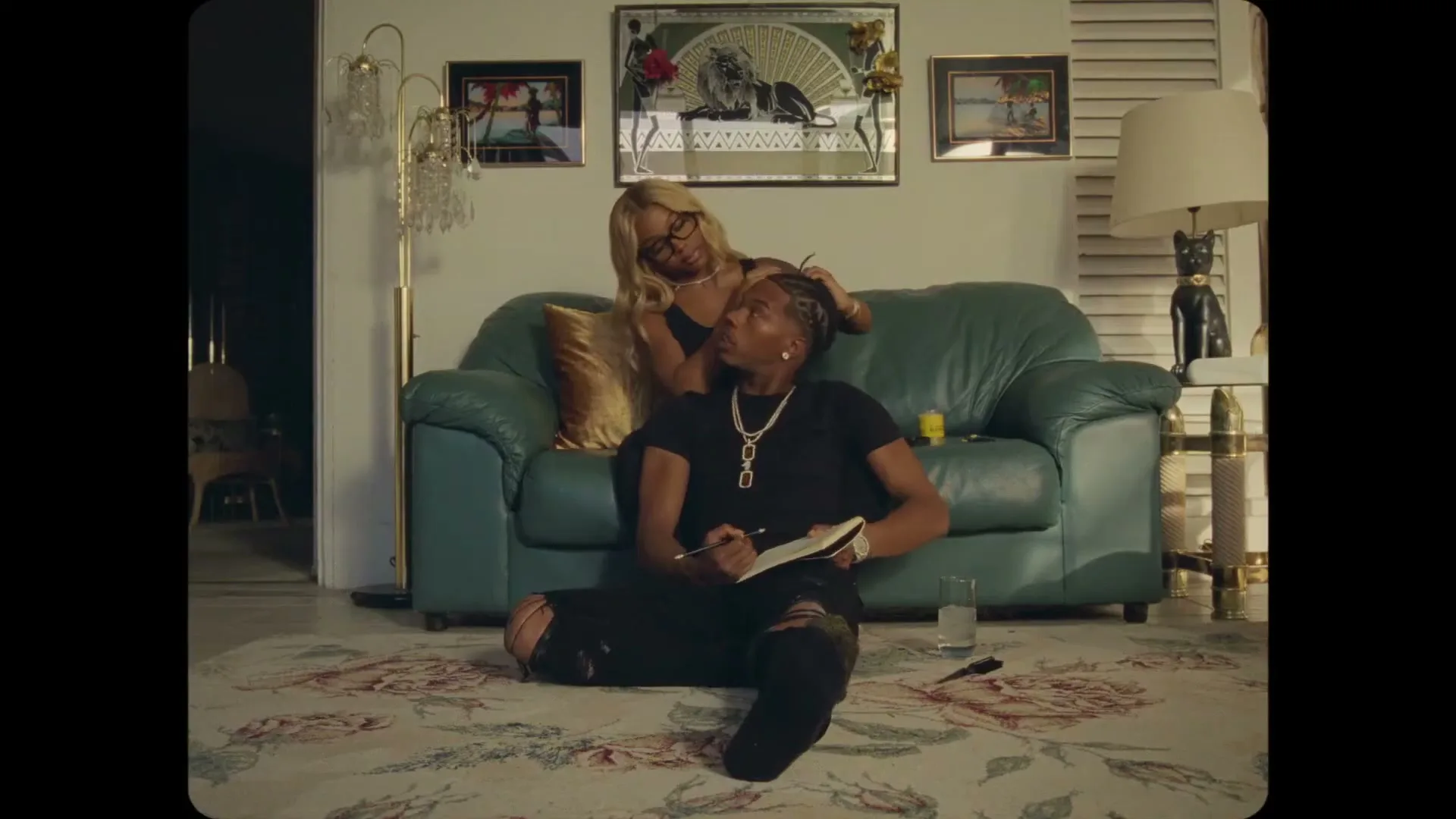A woman with blonde hair sits on a green couch, styling the hair of a man sitting on the floor, who is writing in a notebook in a living room.