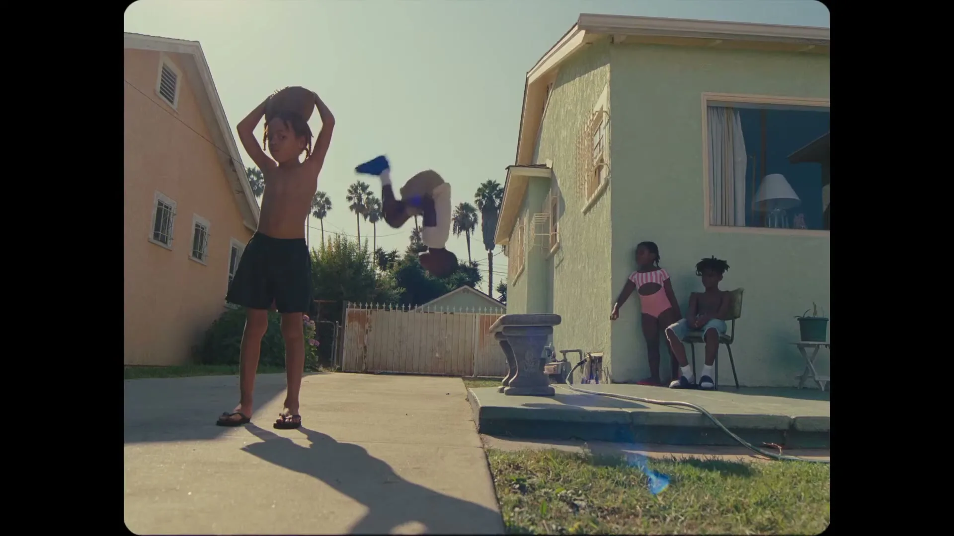 A young boy in shorts does a backflip in a driveway while another boy holds a basketball and two children sit on a porch in a suburban neighborhood.