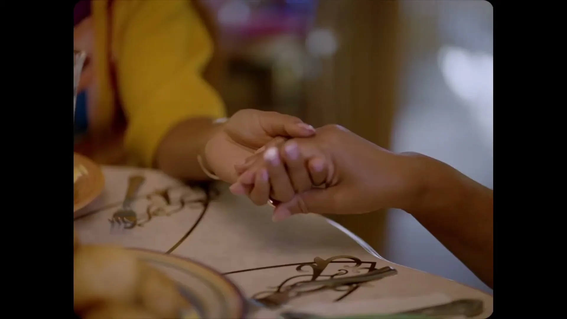 Two people, one wearing a yellow shirt, hold hands across a table with plates and cutlery, showing a moment of connection indoors.