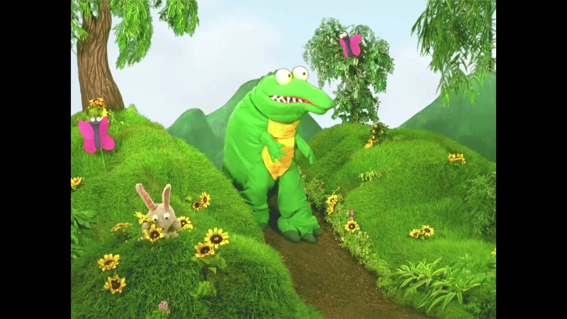A green crocodile puppet with big eyes walks along a path in a vibrant, grassy landscape with a rabbit and butterflies.