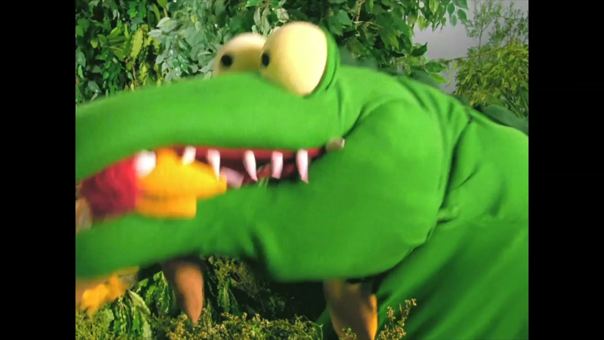 A green crocodile puppet with big eyes eats a red and yellow bird puppet in a jungle setting, with blood splattering from its mouth.