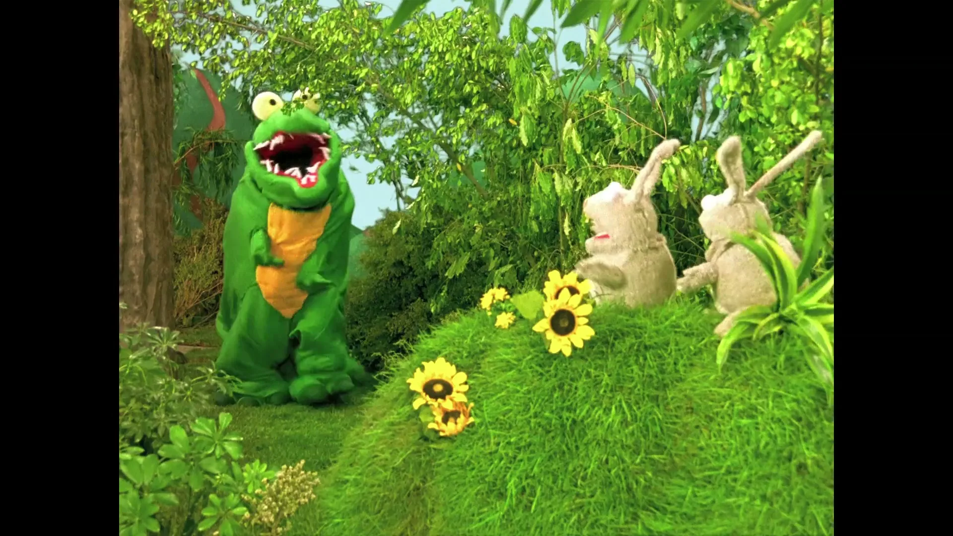 A green crocodile puppet with big eyes and an open mouth stands in a lush green forest, while two rabbit puppets watch from a grassy hill with sunflowers.