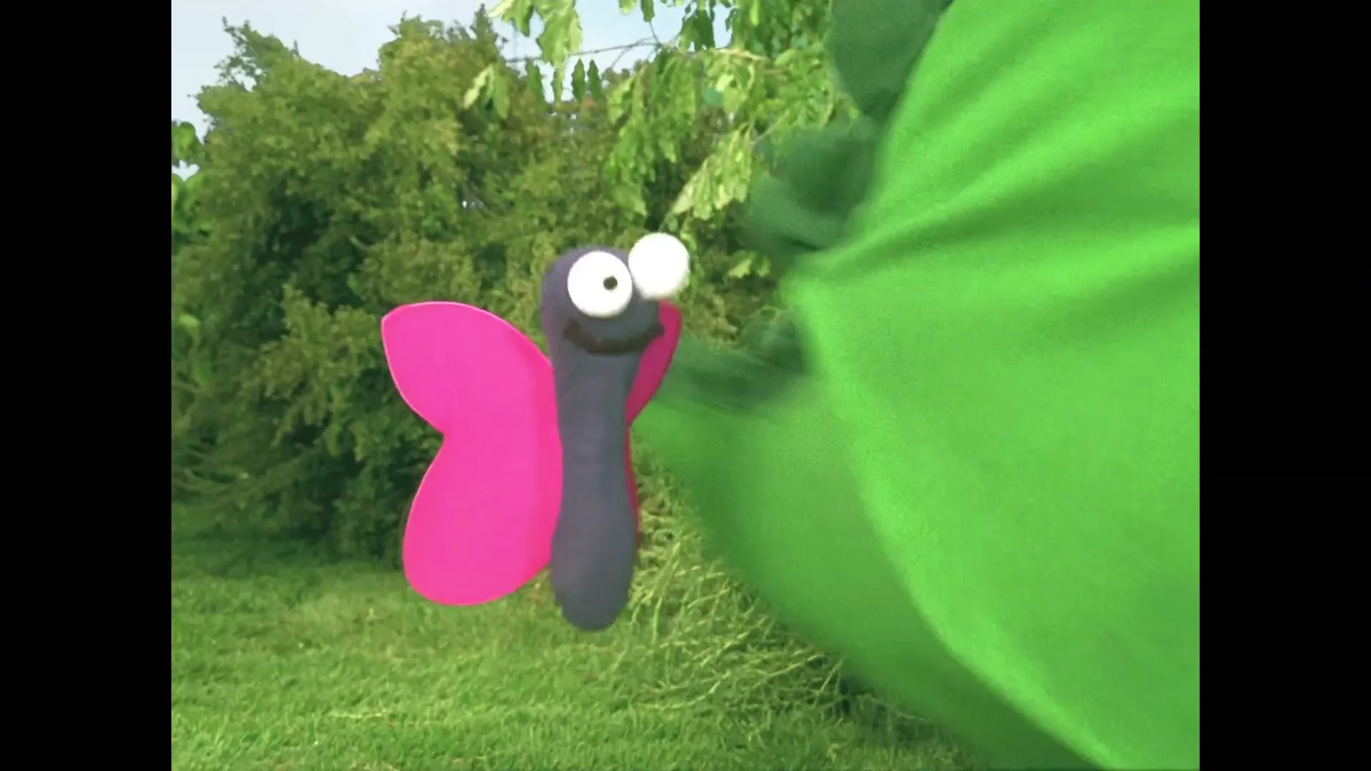 A purple butterfly puppet with pink wings and googly eyes flies in a garden, then is eaten by a green monster puppet.