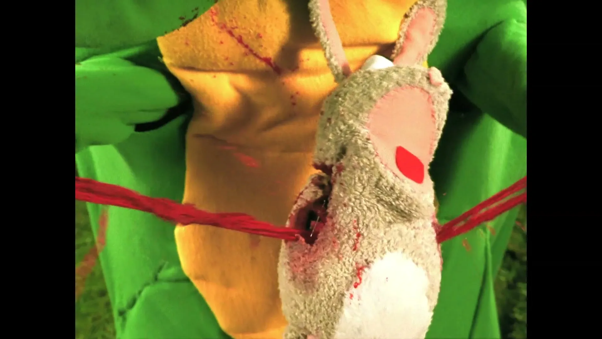 A person in a green and yellow costume holds a grey stuffed bunny, which is then impaled by red strings, splattering blood on the costume.