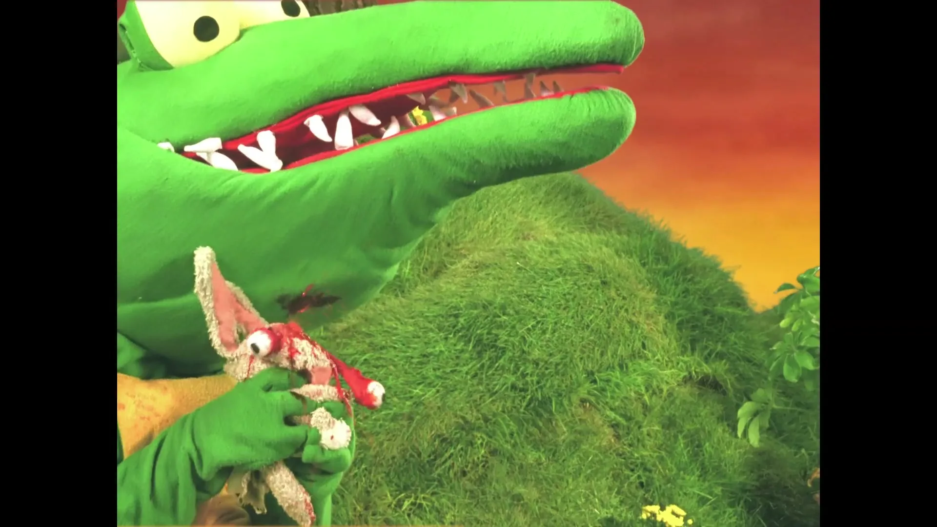 A green crocodile puppet with large white teeth holds a small, bloodied rabbit puppet in its hand, against a backdrop of green hills and an orange sky.