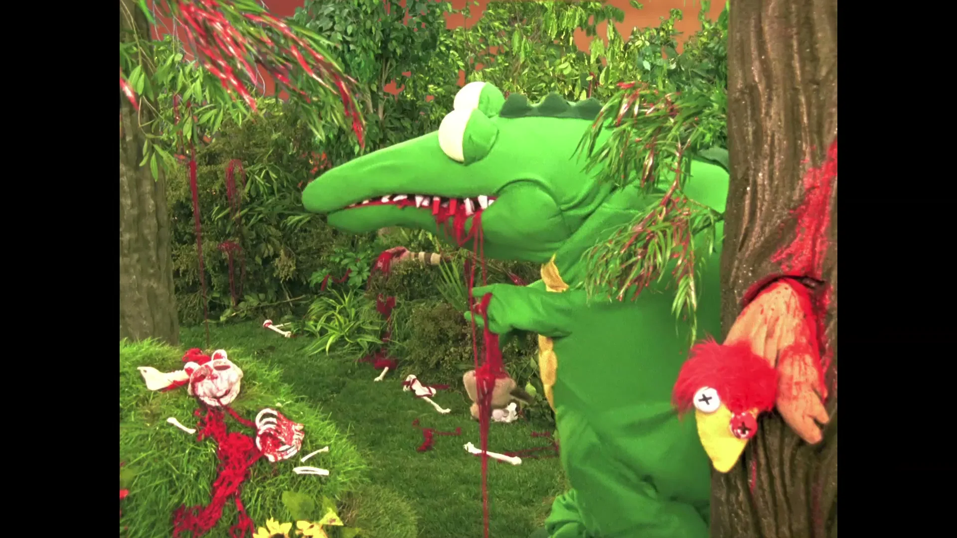 A green crocodile puppet with blood dripping from its mouth stands in a jungle set, surrounded by bones and blood, as a red bird puppet peeks from behind a tree.