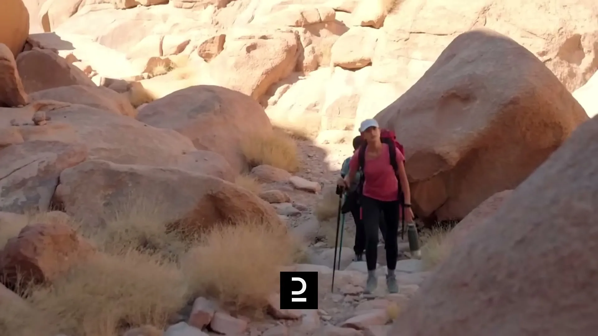 Two women in hiking gear walk through a rocky desert canyon on a sunny day.