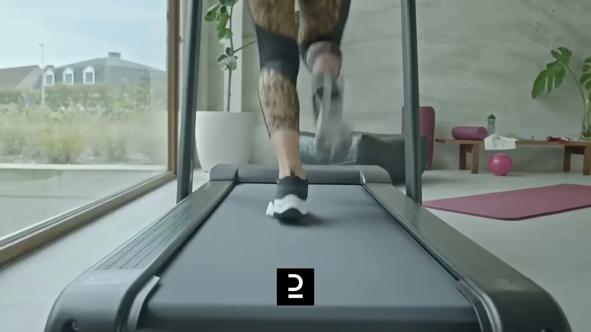 A person in leopard print leggings and black and white sneakers runs on a treadmill in a modern, bright room with large windows.