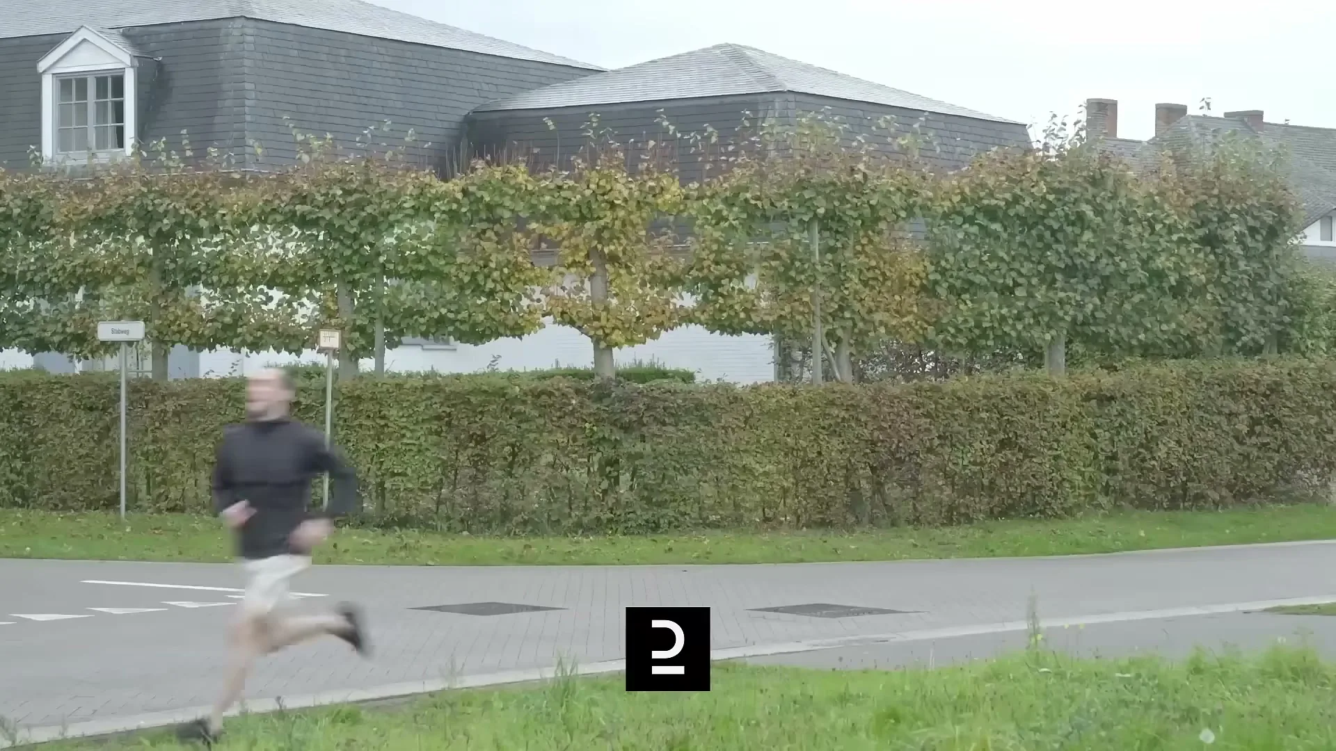 A man in a black hoodie and light shorts runs past a hedge and a house on a suburban street during the day.