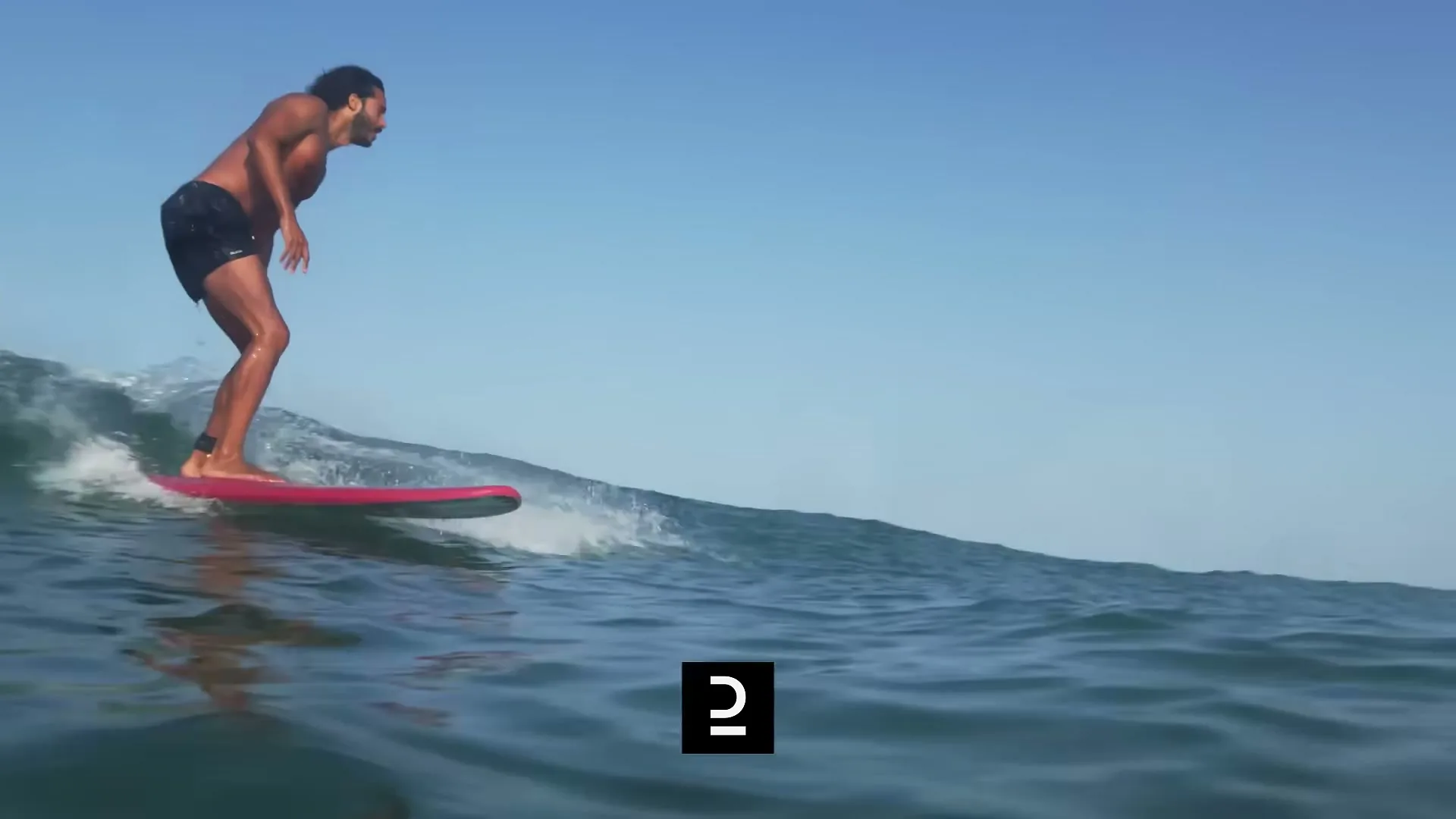 A man with dark hair and a beard surfs on a red surfboard, riding a wave in the ocean under a clear blue sky.