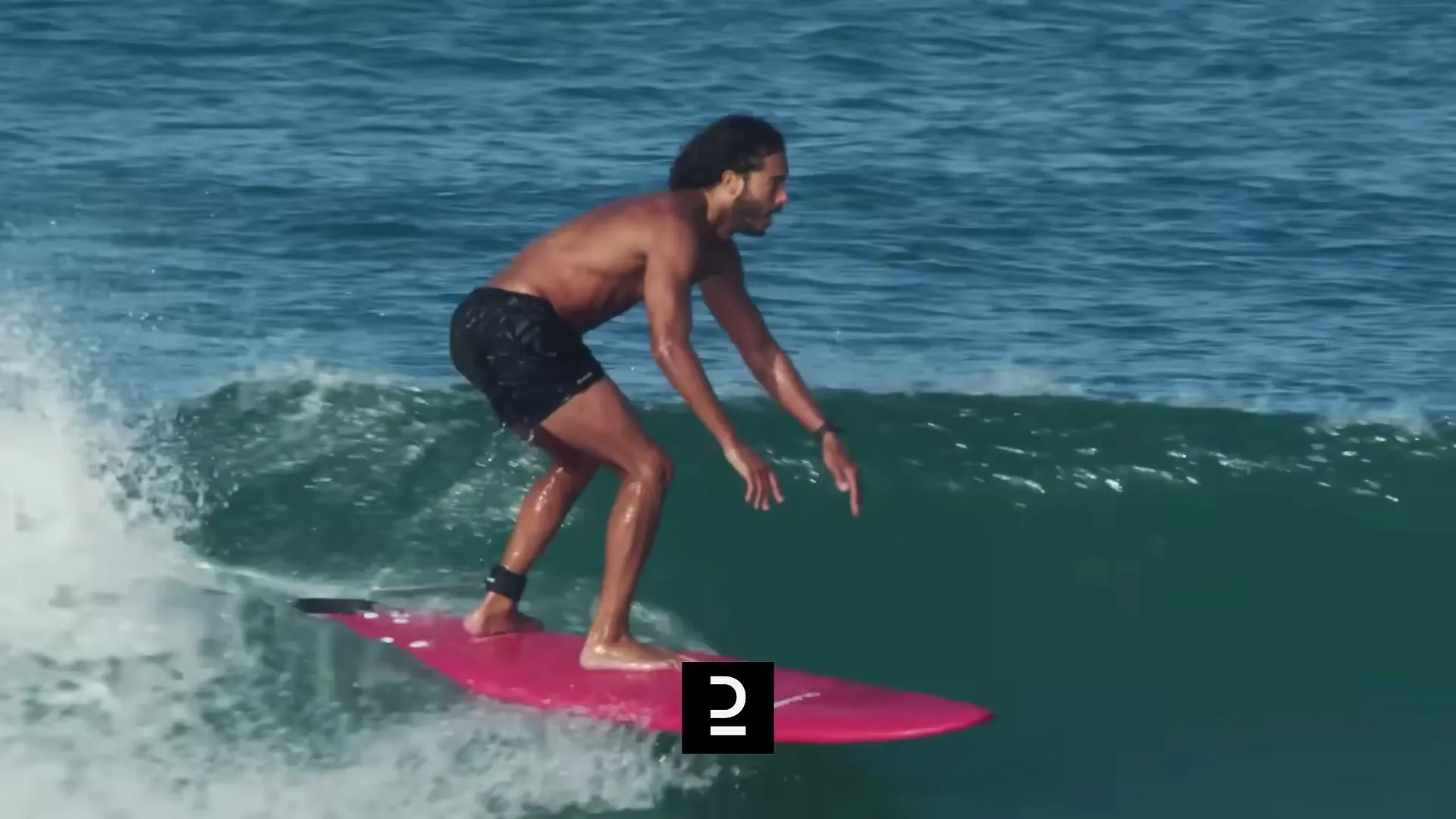 A shirtless man with dark curly hair surfs on a pink surfboard, riding a blue ocean wave under bright daylight.