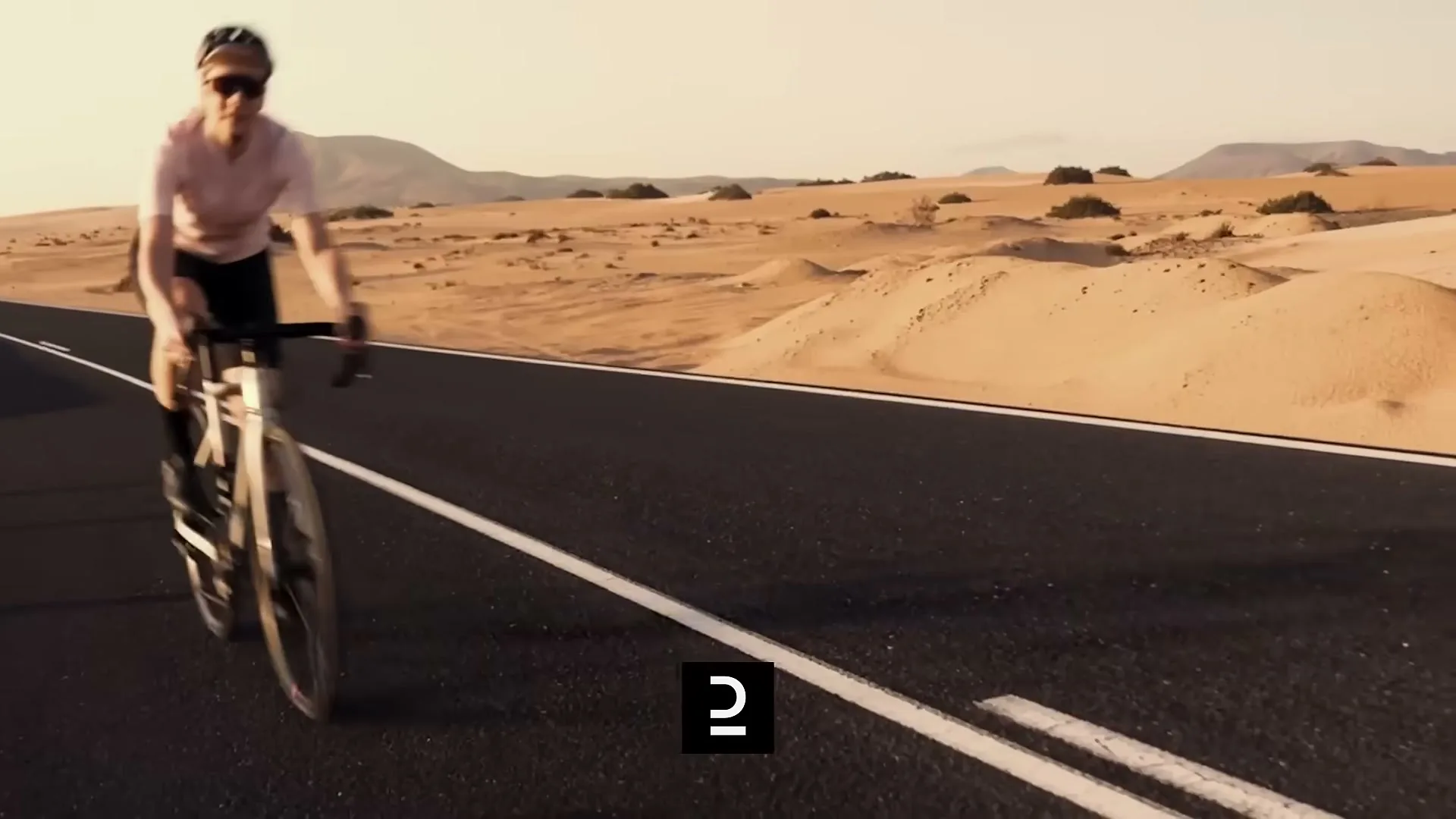 A female cyclist in a pink shirt and black shorts rides a road bike on an empty desert road with sand dunes and mountains in the background.