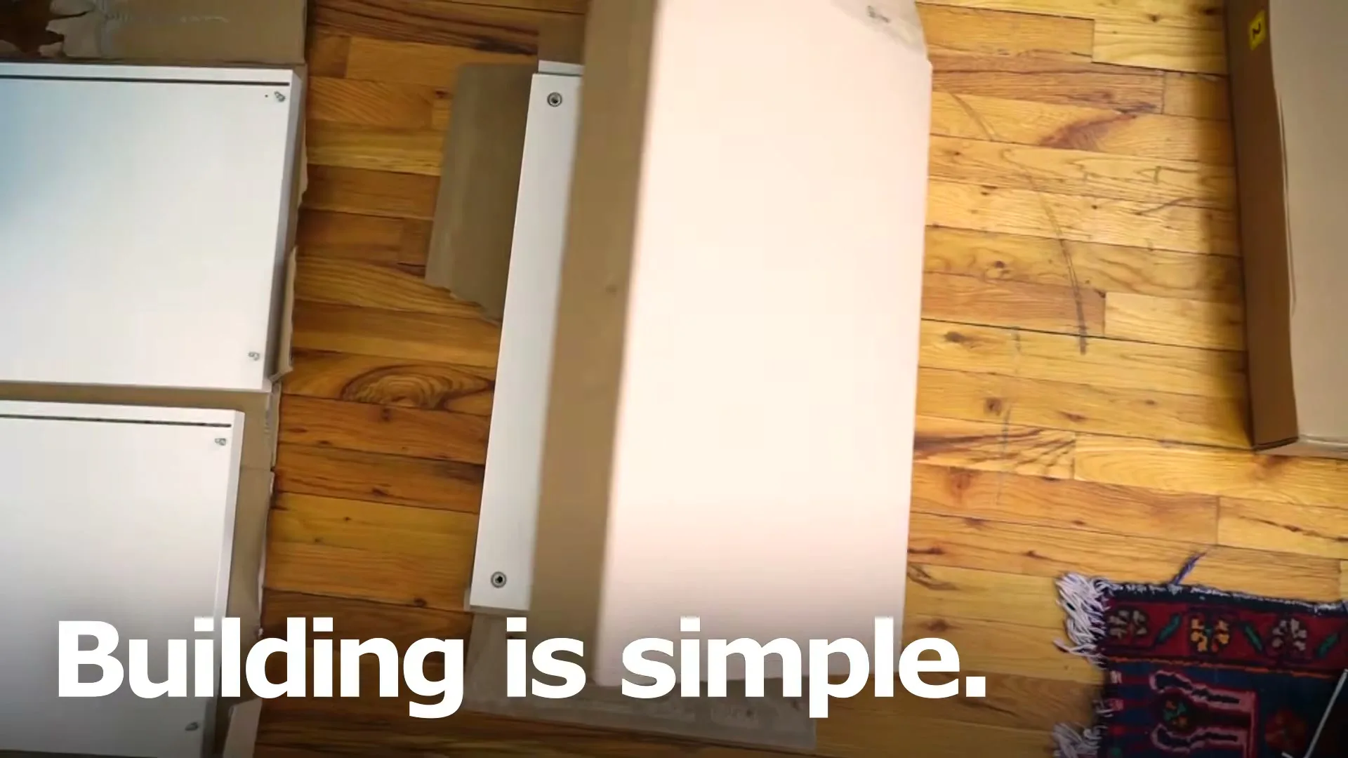 A person's hands assemble a white furniture panel from a cardboard box on a wooden floor, with other panels nearby.