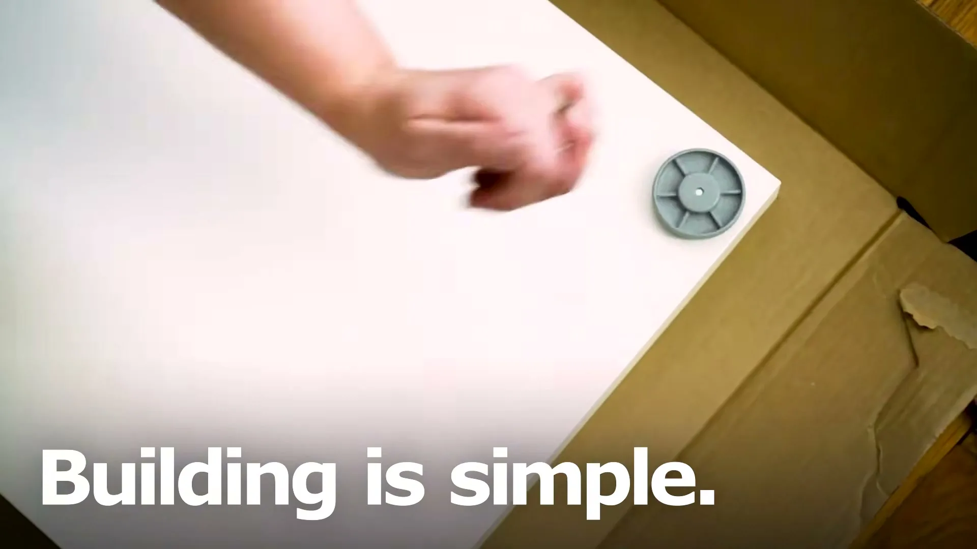 A person's hand places a grey plastic furniture connector onto a white board, inside a cardboard box.