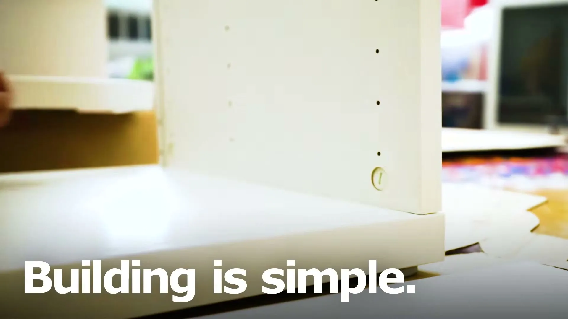 A person's hands assemble a white piece of furniture with a screwdriver, with the text "Building is simple" overlaid.