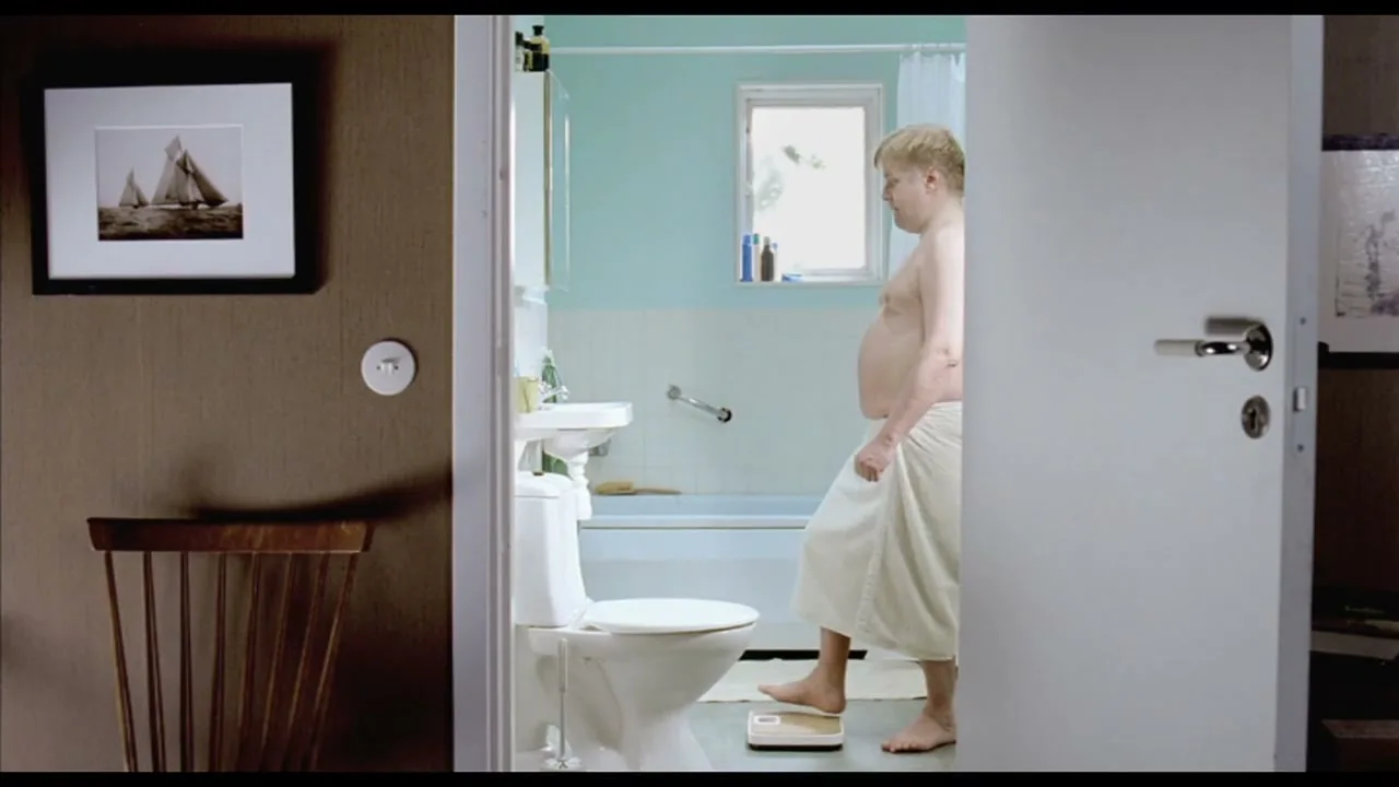 A man in his 30s, wearing a towel, steps onto a scale in a bathroom, looking down at the reading with a concerned expression.
