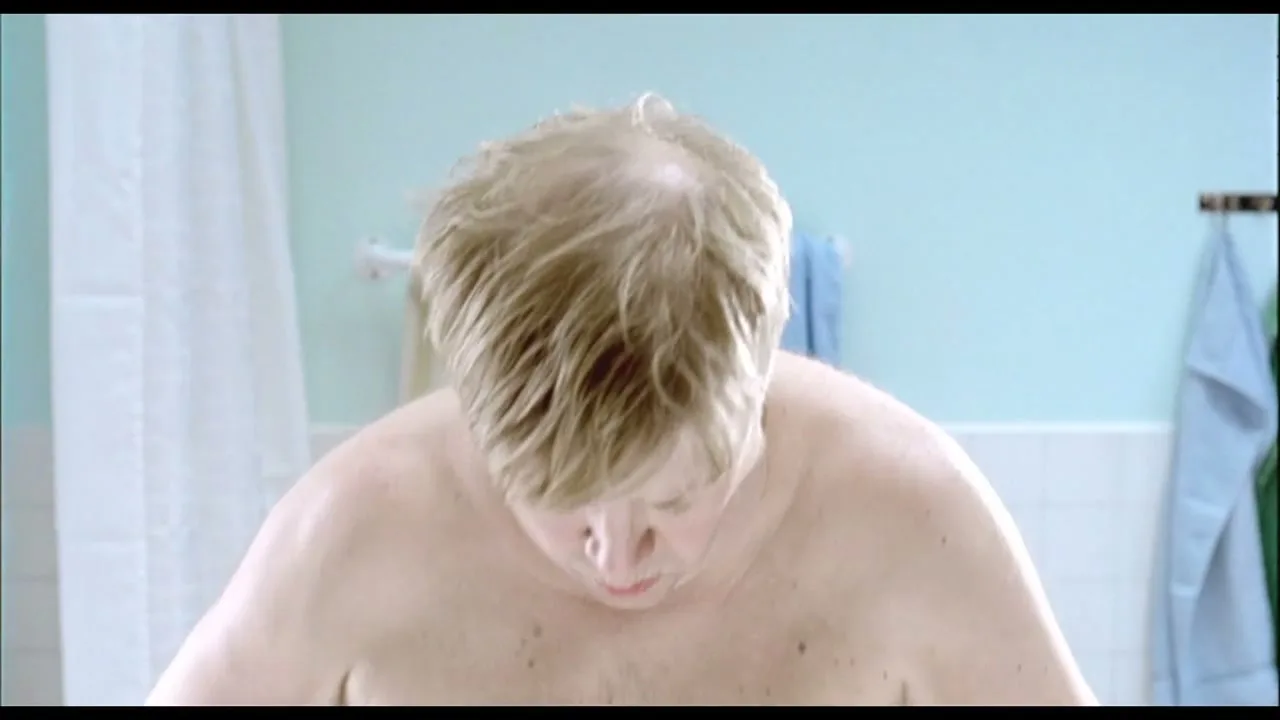 A shirtless, overweight man with blonde hair looks down at a scale, then looks up with a worried expression in a bathroom.