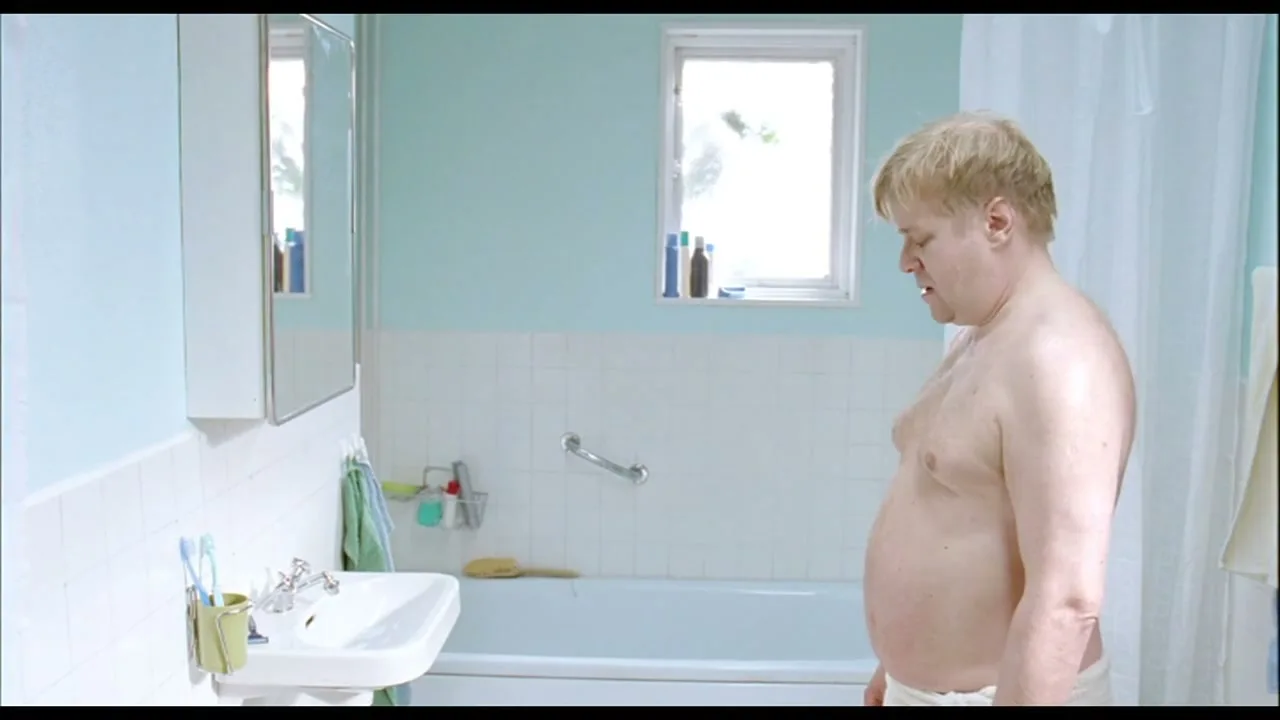 A shirtless, overweight man with blonde hair stands in a bathroom, looking down at a scale off-screen, appearing disappointed.