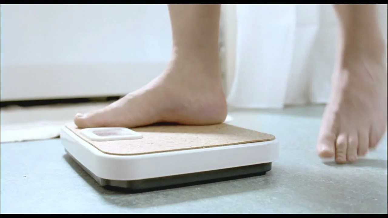 A person's bare feet step onto a bathroom scale, revealing the scale's design and the person's legs in a bright, clean bathroom setting.