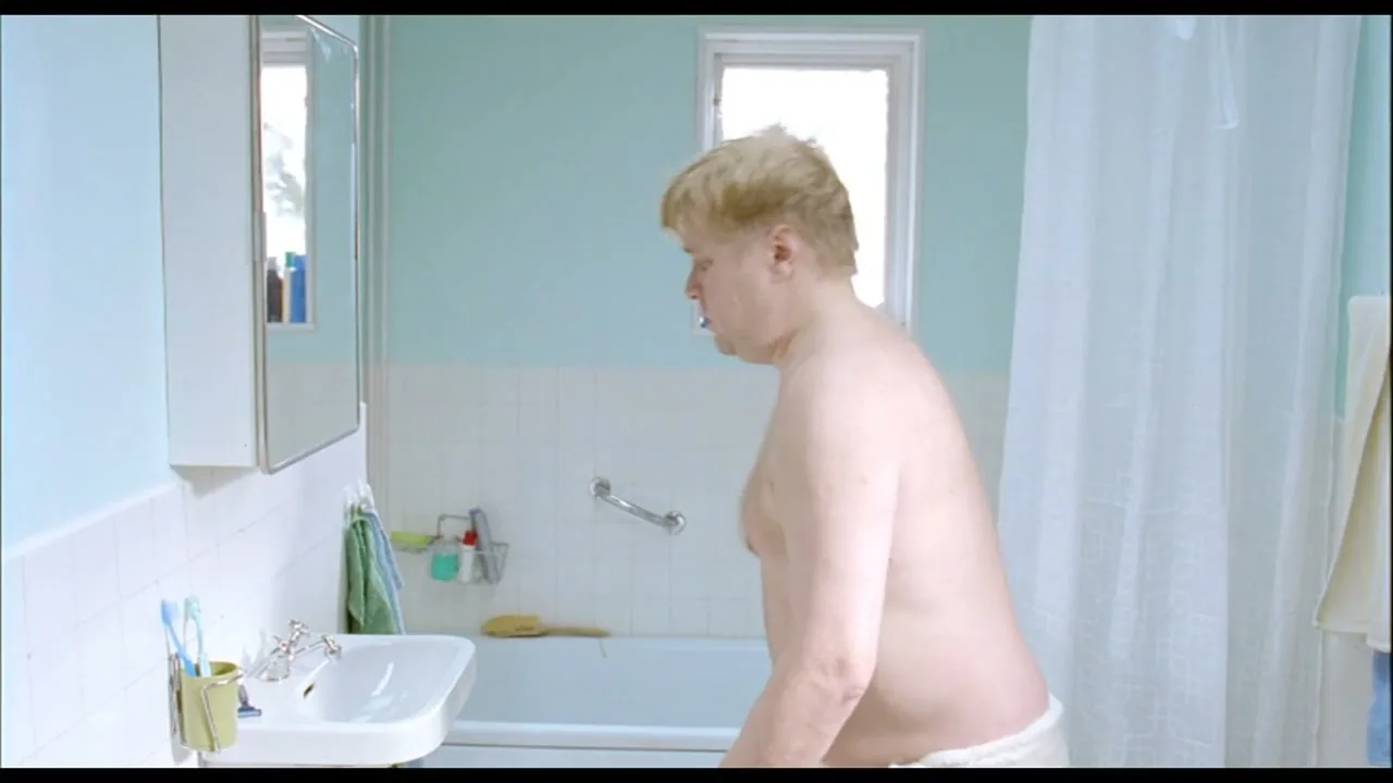 A man with blonde hair, wearing a white towel, stands in a light blue and white bathroom, looking down at his stomach.