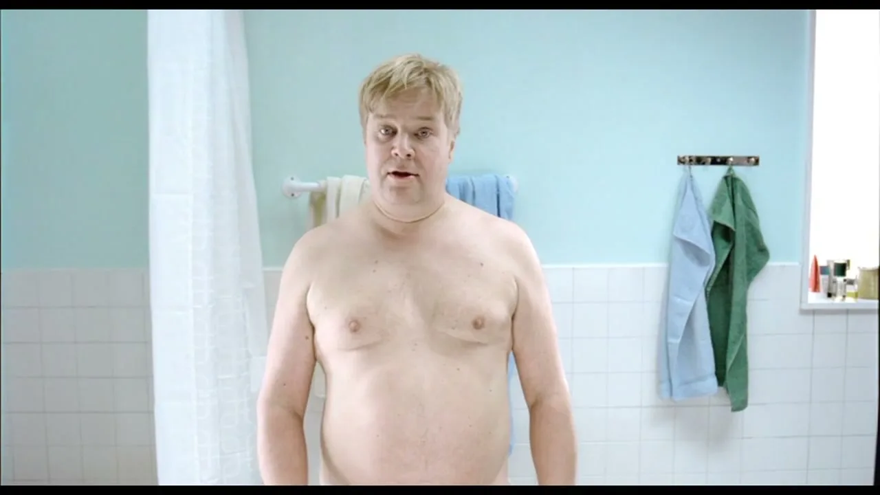 A shirtless, overweight man with blonde hair stands in a bathroom, looking down at a scale, then up at the camera, then down again.