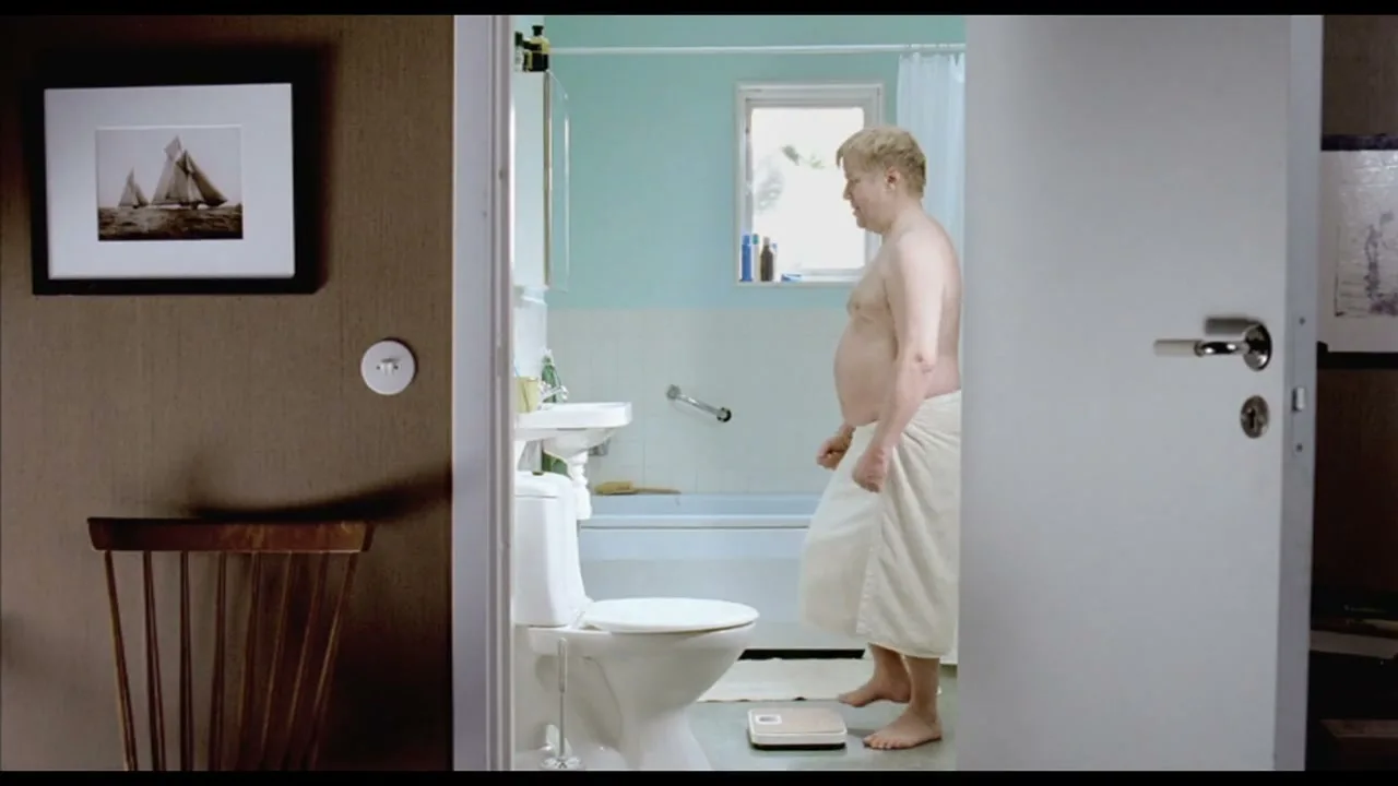 A man in his 30s, wearing a towel, stands on a scale in a bathroom, looking down at the reading with a disappointed expression.
