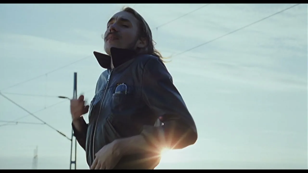 A man with long hair and a mustache, wearing a dark jacket, runs with a determined expression against a bright sky with power lines.