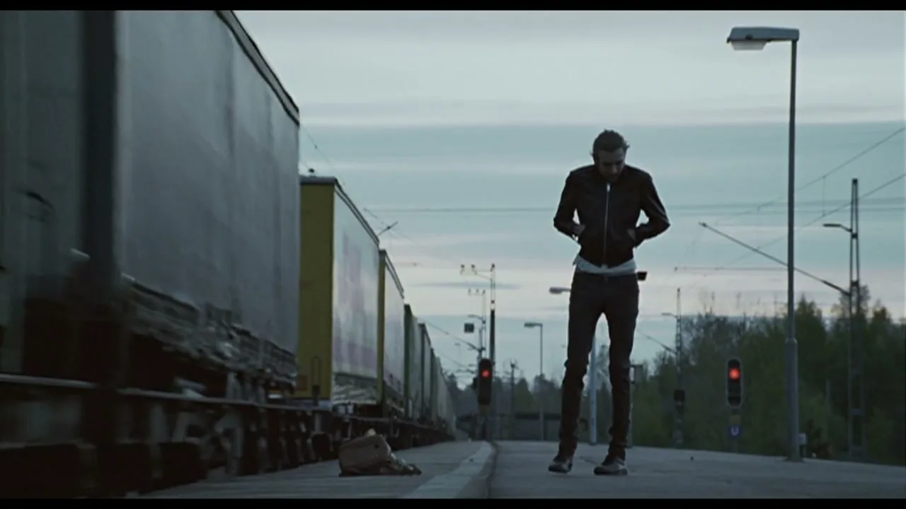 A man in a black leather jacket stands on a train platform as a freight train passes by, looking down at a bag on the ground.