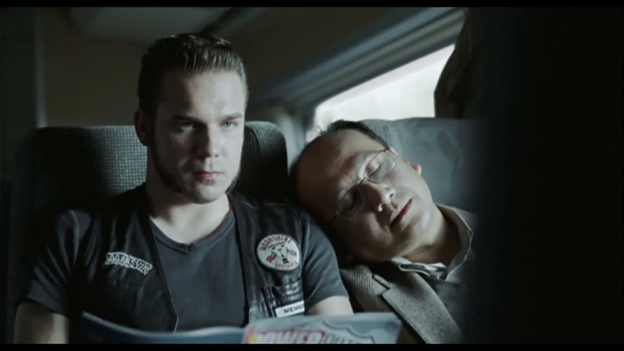 A young man in a biker vest reads a magazine next to an older man sleeping on a train.