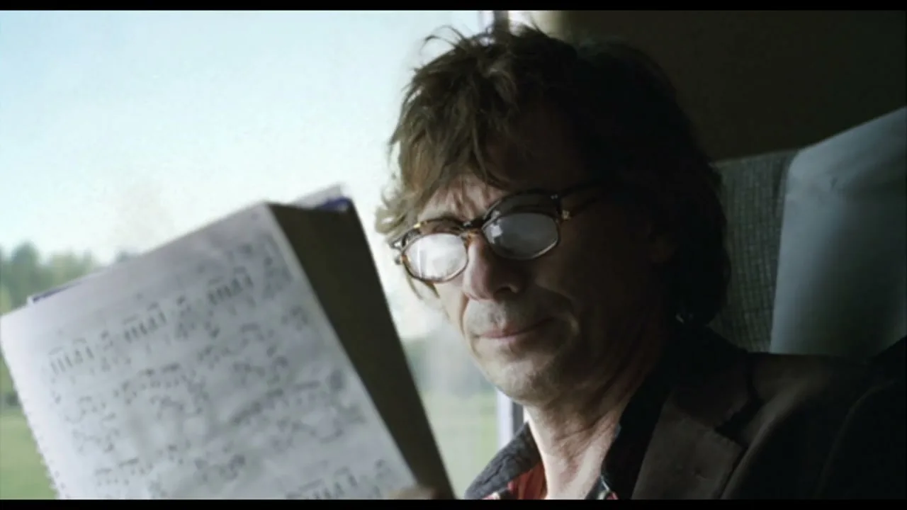 A man with messy hair and glasses reads sheet music on a train, looking thoughtful.