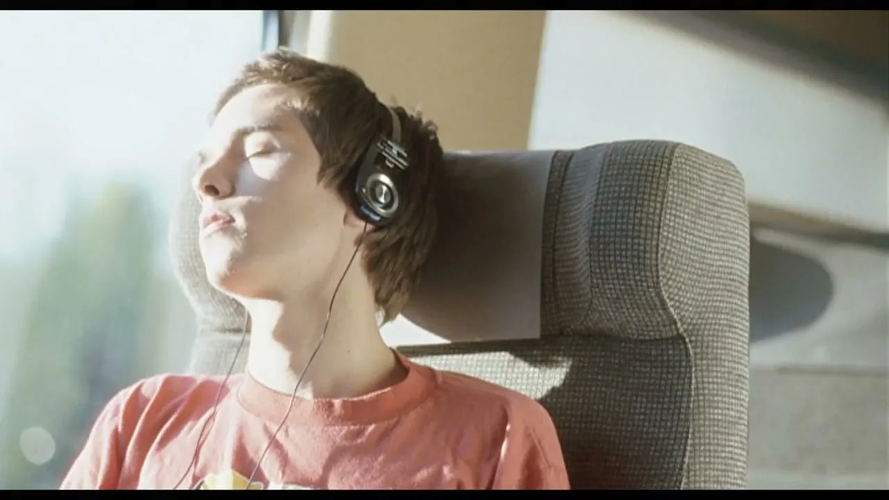 A young man with headphones on is sleeping in a train seat, with sunlight on his face.