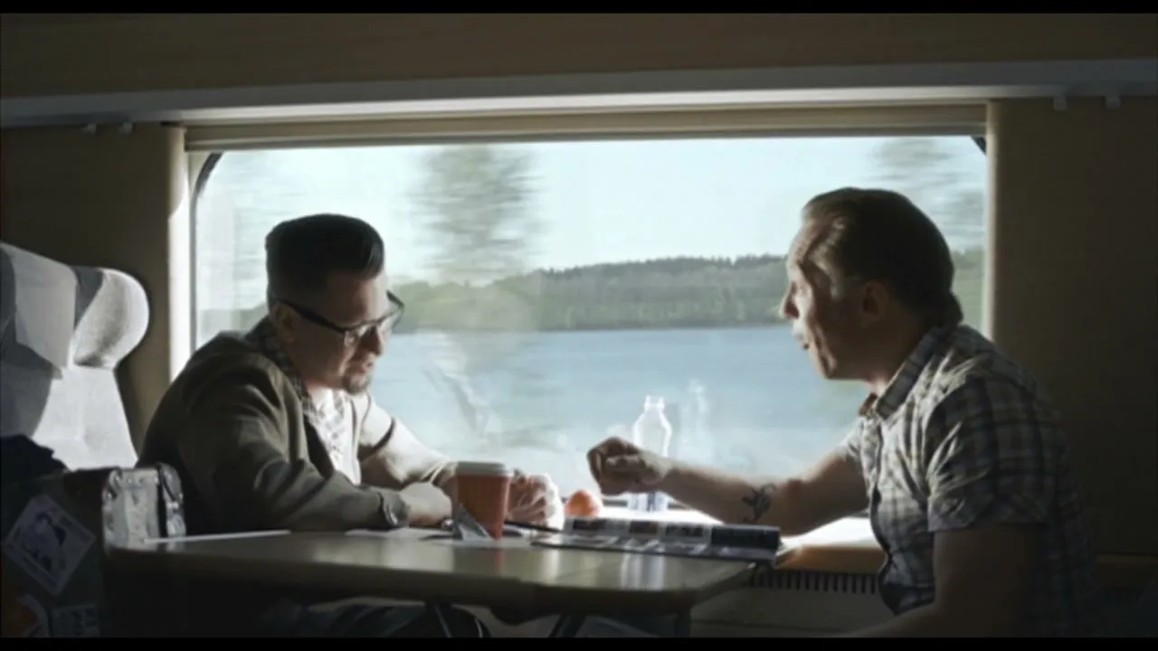 Two men sit opposite each other at a table on a train, talking, with a lake and trees visible outside the window.
