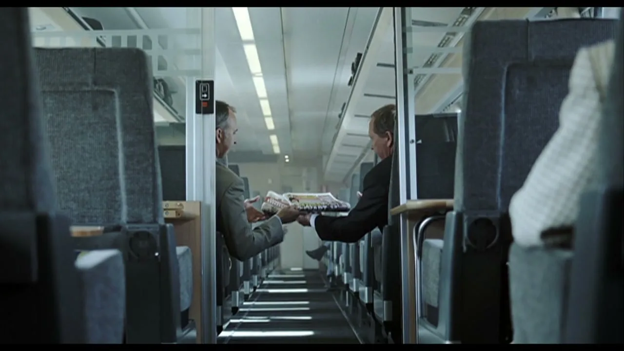 Two men in suits on a train pass a magazine to each other, seen from between seats in the aisle.
