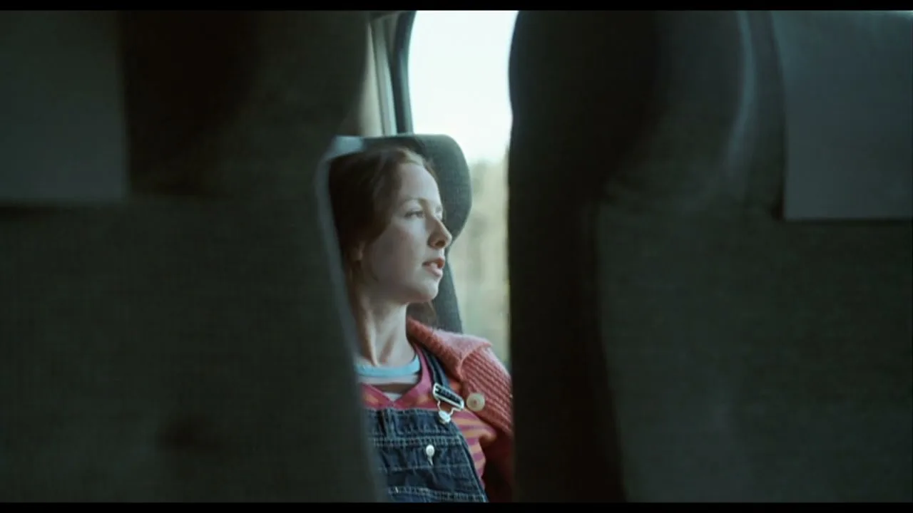 A young woman with red hair, wearing overalls and a pink cardigan, looks out the window of a moving train, framed by two seats.