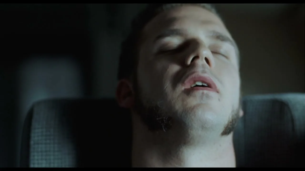 A young man with sideburns is seen in a medium closeup, eyes closed, head tilted back, appearing to be asleep or unconscious in a dark interior.