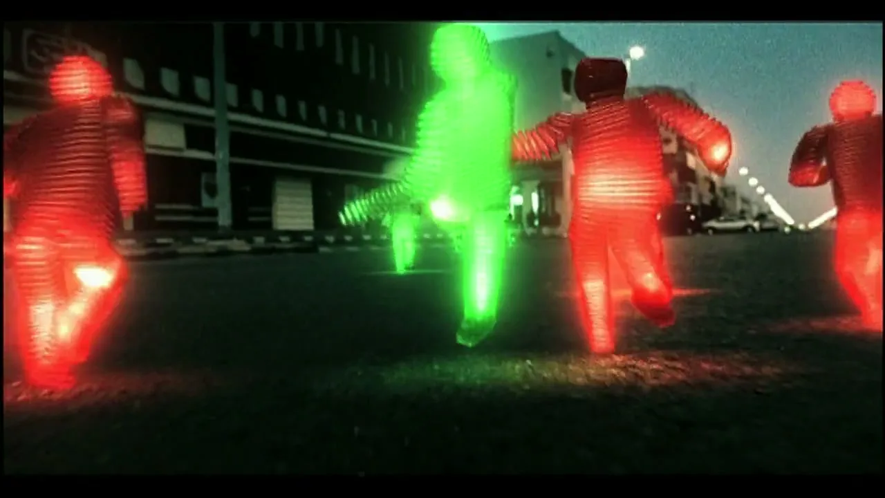 A group of glowing, blocky figures, one green and others red, run down a street at dusk, with buildings in the background.