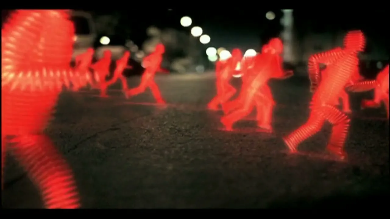 Multiple glowing red, translucent, human-shaped figures run across a dark, cracked asphalt surface at night, with blurred streetlights and vehicles in the background.