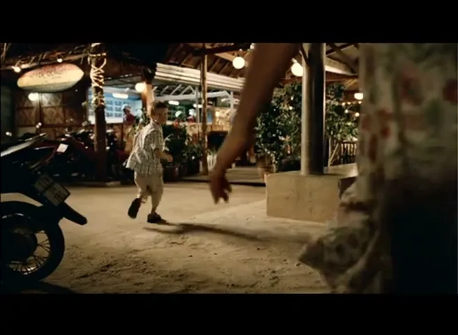 A young boy runs towards the camera at night, then two adults walk past him, in front of a restaurant.