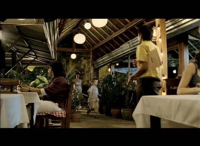 A young boy in a plaid shirt walks through an outdoor restaurant, with a woman and a waiter in the foreground and background.