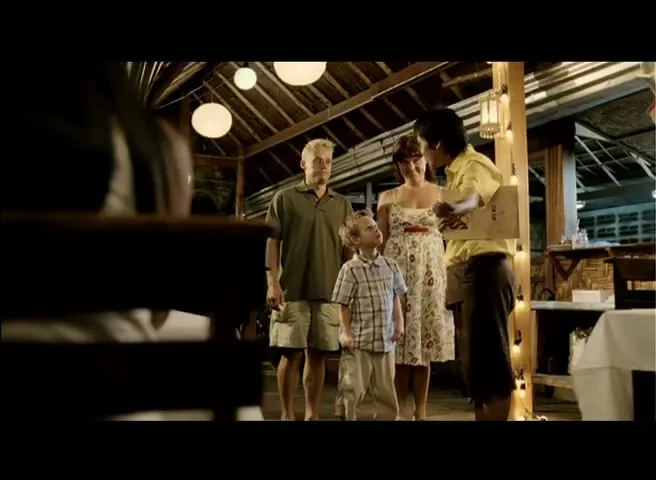 A family of three, a blonde man, a woman in a floral dress, and a young boy, stand barefoot in a dimly lit, open-air restaurant, looking at a male waiter in a yellow shirt.