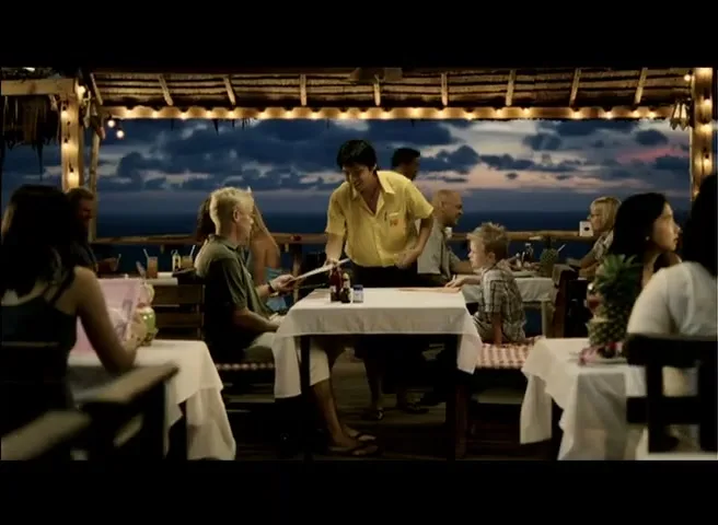 A waiter in a yellow shirt serves a family with a young boy at an outdoor restaurant with a view of the ocean at dusk.